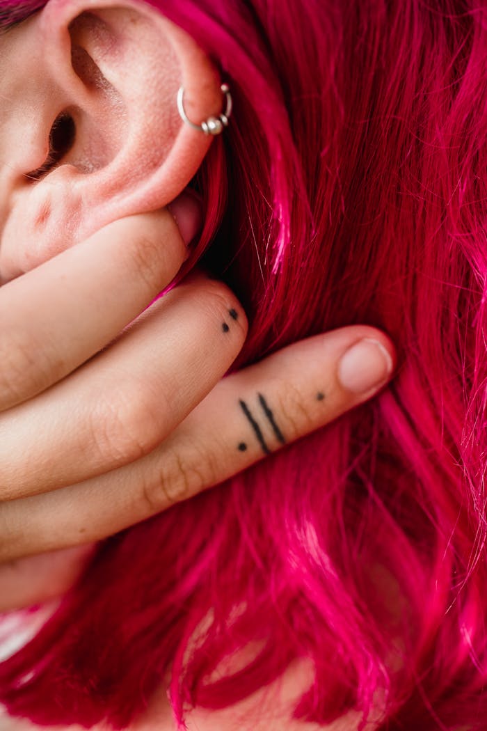 Close-up of a woman's hand with tattoos touching vibrant pink hair near her ear.